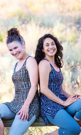 Two women sitting back-to-back outdoors, wearing matching blue and white checkered dresses.