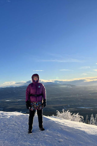 Person standing on a snowy mountain top with a scenic view of mountains and clouds.